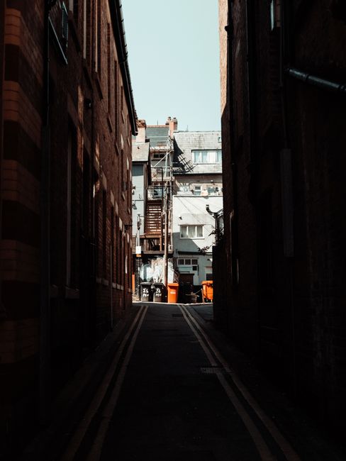 A black and white photograph of a narrow street in a residential area during daytime, showing brick apartment buildings on both sides. The left side features a street sign reading 'Meadowcroft Mews' a