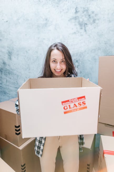 A young woman with long brown hair smiling at camera while holding an empty cardboard moving box labeled 'GLASS' in a room with light blue textured walls. Several other packed cardboard boxes, some with black and red tape, are stacked on the floor around her. The boxes are made of brown corrugated cardboard and vary in size, indicating a packing process during home relocation. The woman is dressed casually in a plaid shirt and light-colored trousers. Behind her, the environment suggests an organised packing stage in a house or flat, typically part of furniture transport and moving logistics. This scene is associated with professional removals services such as those offered by Man and Van Brent, highlighting packing and moving procedures in a domestic setting.
