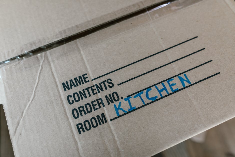 Close-up view of a cardboard moving box with printed labels and handwritten notes, specifically indicating the contents as kitchen items. The box features black printed lines for writing details such as name, contents, order number, and room, with the word 'KITCHEN' handwritten in blue ink next to the 'ROOM' label. The box is partially sealed with clear packaging tape, and it appears to be ready for or during the process of home relocation. It is situated on or near other packing materials, possibly inside an interior space preparing for furniture transport or packing and moving activities, as handled by a professional removals service like Man and Van Brent, which offers house removals and relocation solutions in Willesden Green.
