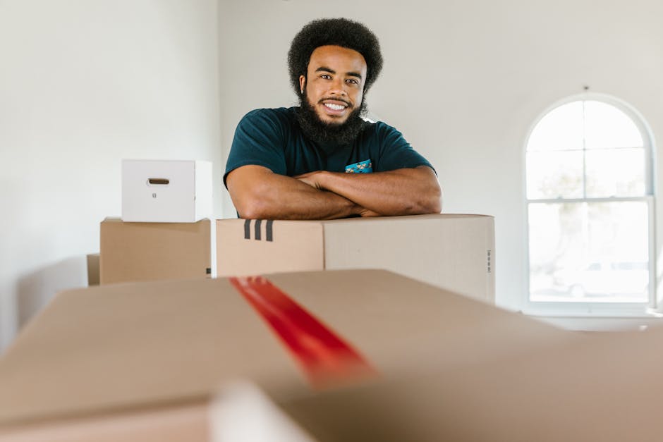 A young woman with long brown hair smiling at camera while holding an empty cardboard moving box labeled 'GLASS' in a room with light blue textured walls. Several other packed cardboard boxes, some with black and red tape, are stacked on the floor around her. The boxes are made of brown corrugated cardboard and vary in size, indicating a packing process during home relocation. The woman is dressed casually in a plaid shirt and light-colored trousers. Behind her, the environment suggests an organised packing stage in a house or flat, typically part of furniture transport and moving logistics. This scene is associated with professional removals services such as those offered by Man and Van Brent, highlighting packing and moving procedures in a domestic setting.